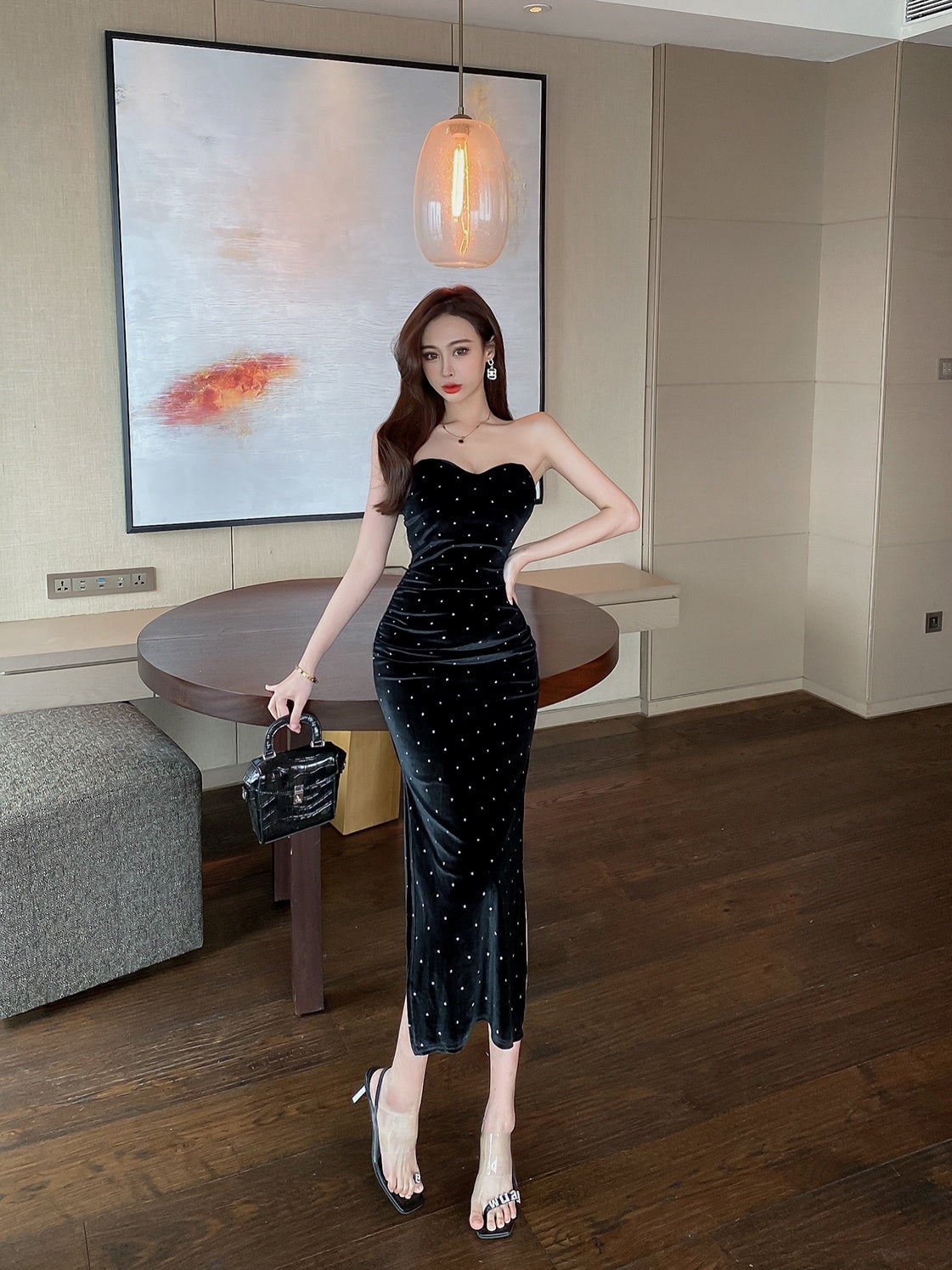 "Look chic et sophistiqué : robe noire moulante à pois, sac à main noir et sandales transparentes, posant devant une décoration moderne.